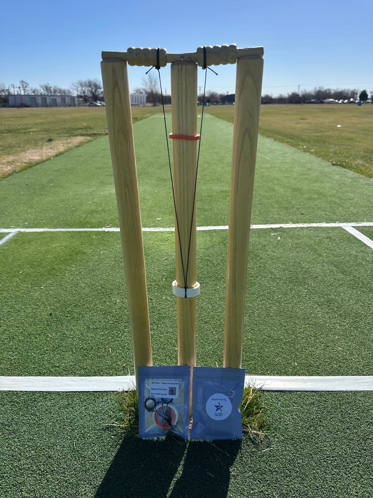 Three stumps on a matting pitch with a bail guard fitted: a black tether runs from each bail down to an orange stopper and a white collar on the middle stump. Two packets rest against the base of the stumps.