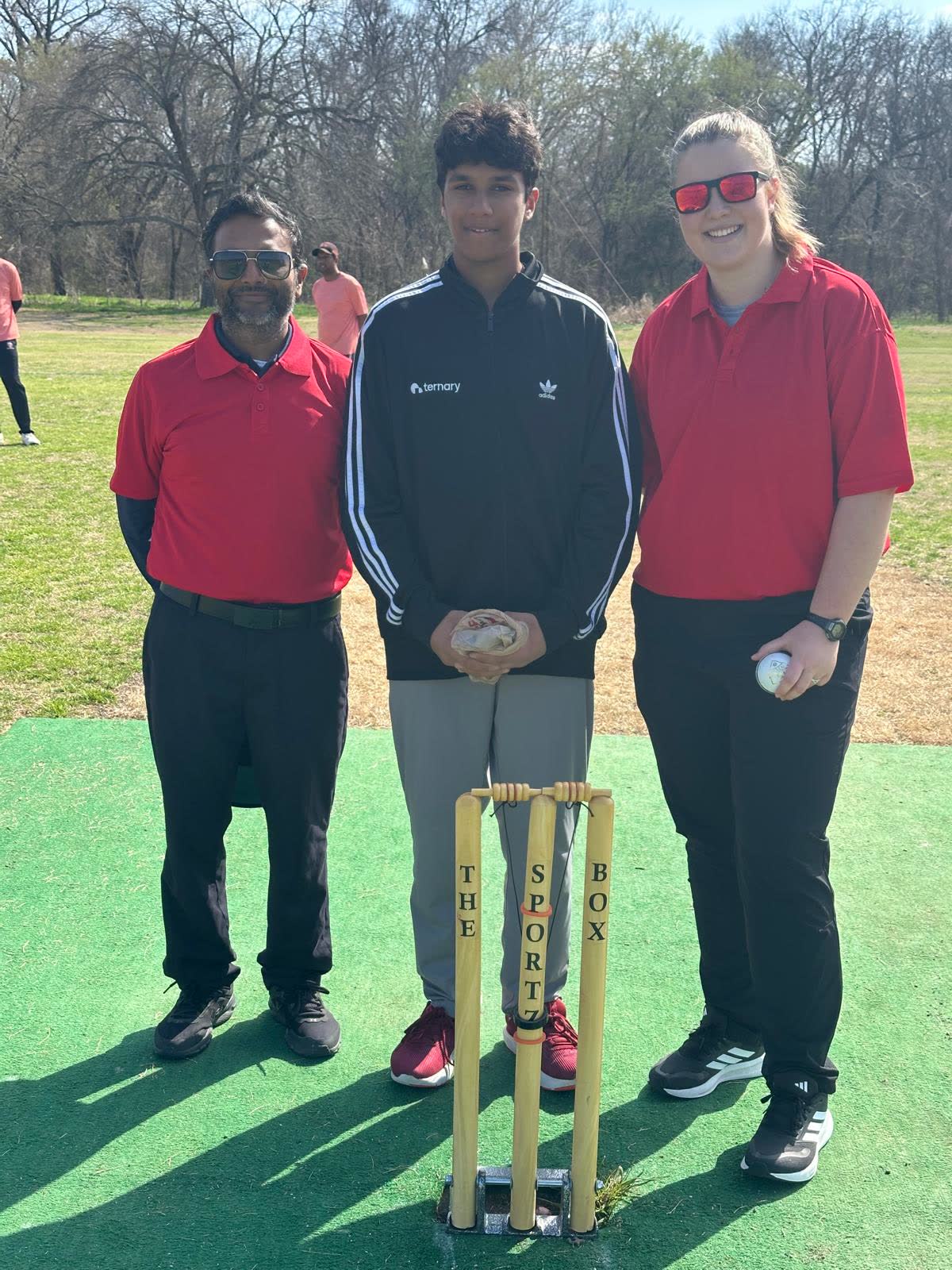 Two umpires in red shirts and a young player standing behind a set of stumps on a matting pitch, the bails tethered by a bail guard.