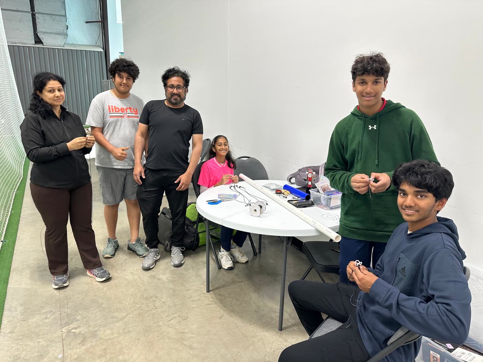 Six volunteers, adults and teenagers, around a folding table in an indoor cricket facility, threading tethers and O-rings by hand.