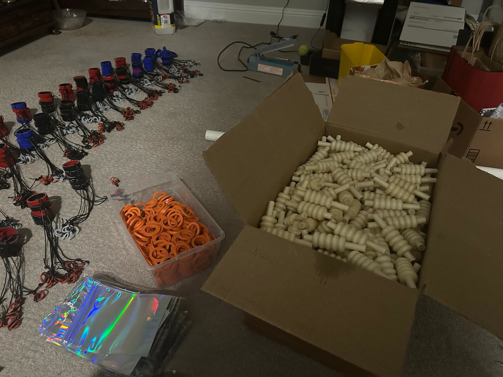 A living room floor covered in bail guard parts: rows of assembled collars and tethers, a tub of orange O-rings, a box of bails, empty packets and a heat sealer.