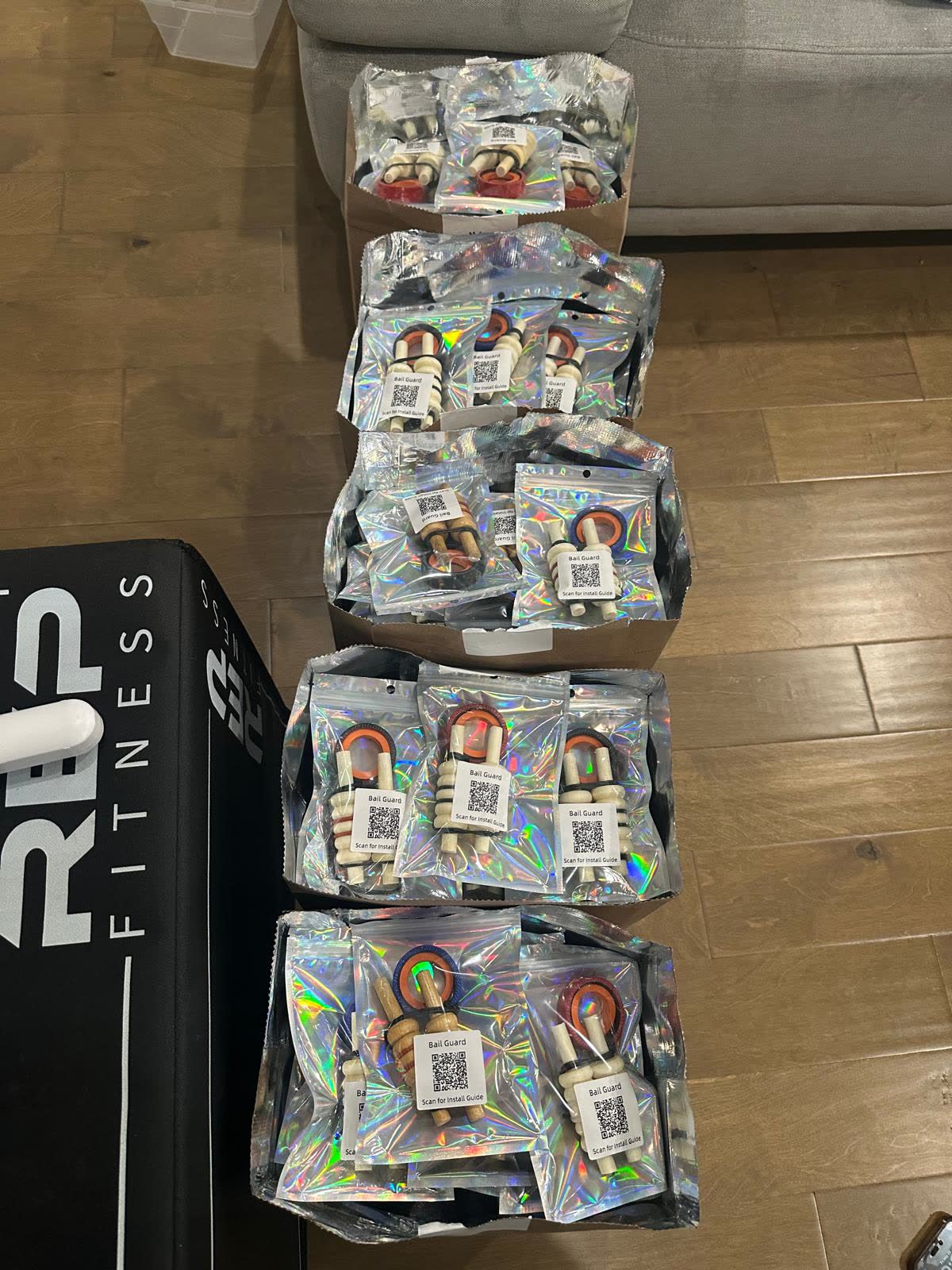 Five open boxes on a wooden floor, each filled with sealed foil packets containing a bail guard and a QR code labelled Scan for install guide.