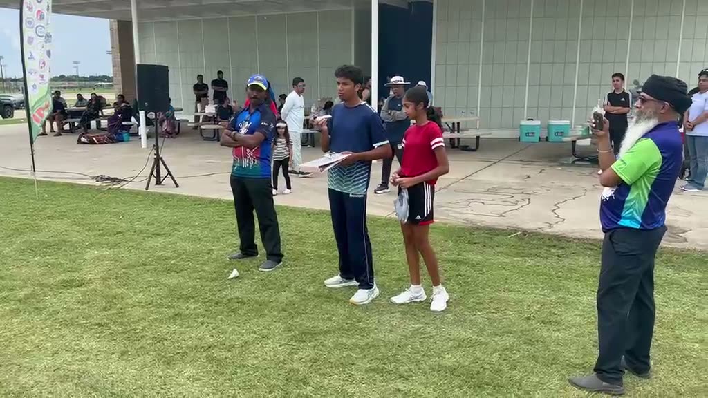 A prize ceremony on the outfield: young players lined up with a microphone, watched by parents and officials under a pavilion.